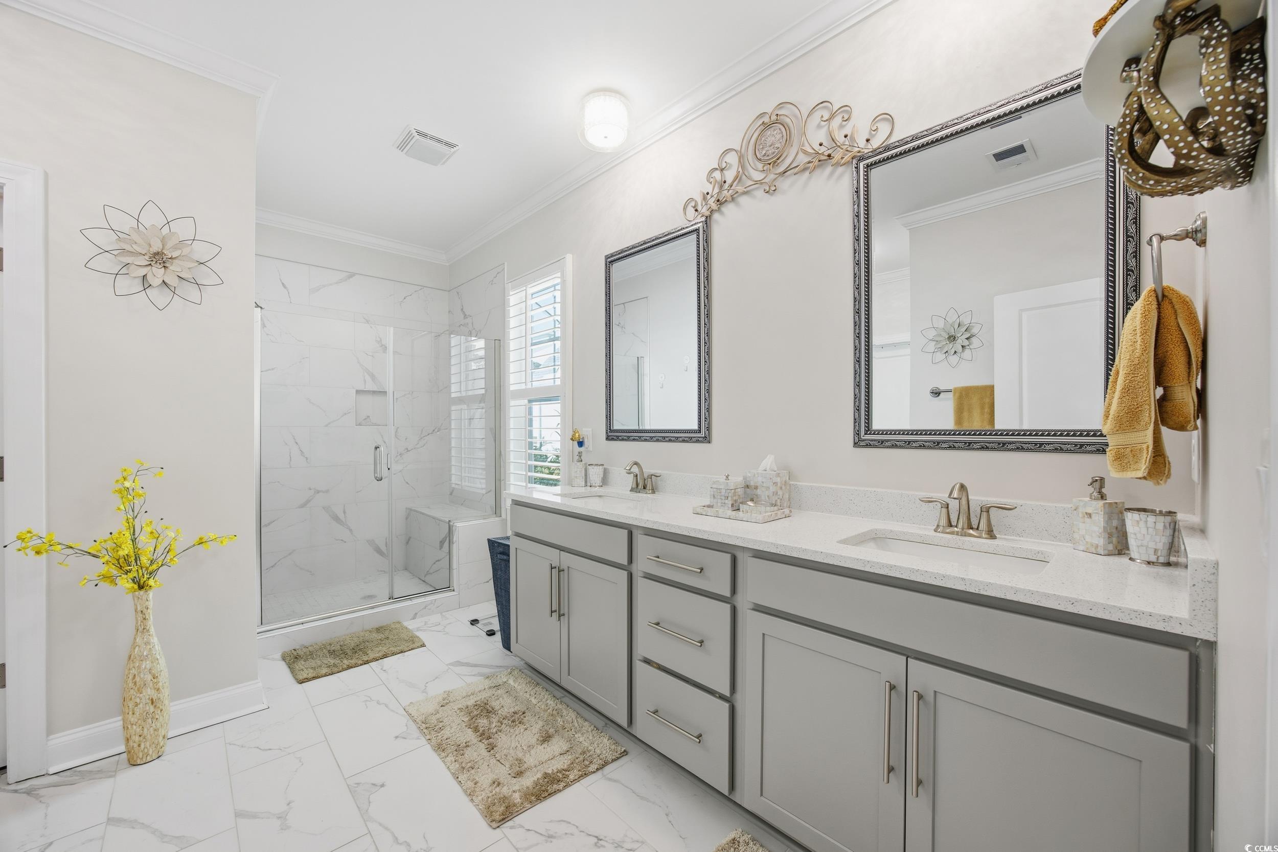 Full bath with crown molding, double vanity, light marble finish floors, and a marble finish shower