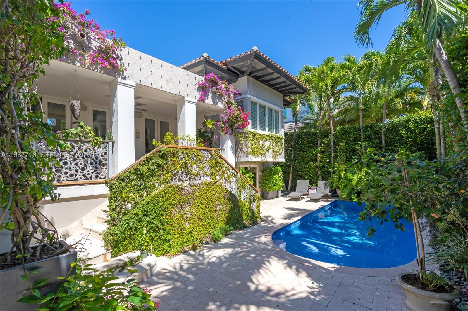 Elevated, 3,888 sf Adjusted Area. Sophisticated Mediterranean beach house, elevated and privately set, offering 5 bedrooms, 5 bathrooms, and an additional powder room, surrounded by a lush, mature backyard garden. A double-height cathedral ceiling in the great room fills the home with natural light and opens to a covered terrace overlooking the pool patio, creating seamless indoor-outdoor living. The great room flows into a warm family room and gourmet kitchen, well suited for everyday living and entertaining, and every bedroom is en suite. The serene primary suite includes a sitting area ideal for relaxing or reading.A spacious den with abundant windows and its own full bath offers flexibility as a home office, guest suite, studio, or cabana, with direct access to the patio and pool. Outdoors, enjoy a private setting with an upper veranda shaded by a living canopy of fig vines, an inviting space for dining and lounging, plus an outdoor gourmet area with a wood-fired pizza oven and tools. Additional highlights include solar panels with backup batteries for energy savings and reliability. Two batteries plus potential for expansion, two garages, and a pool with refreshed tile and grout for a crisp, updated finish. Walking distance to the beach, parks, dining, shopping, and everyday conveniences, with convenient access to Miami. Not Included in the sale: all display shelving, two bronze candle holders, and the chandelier hanging above the stairs.