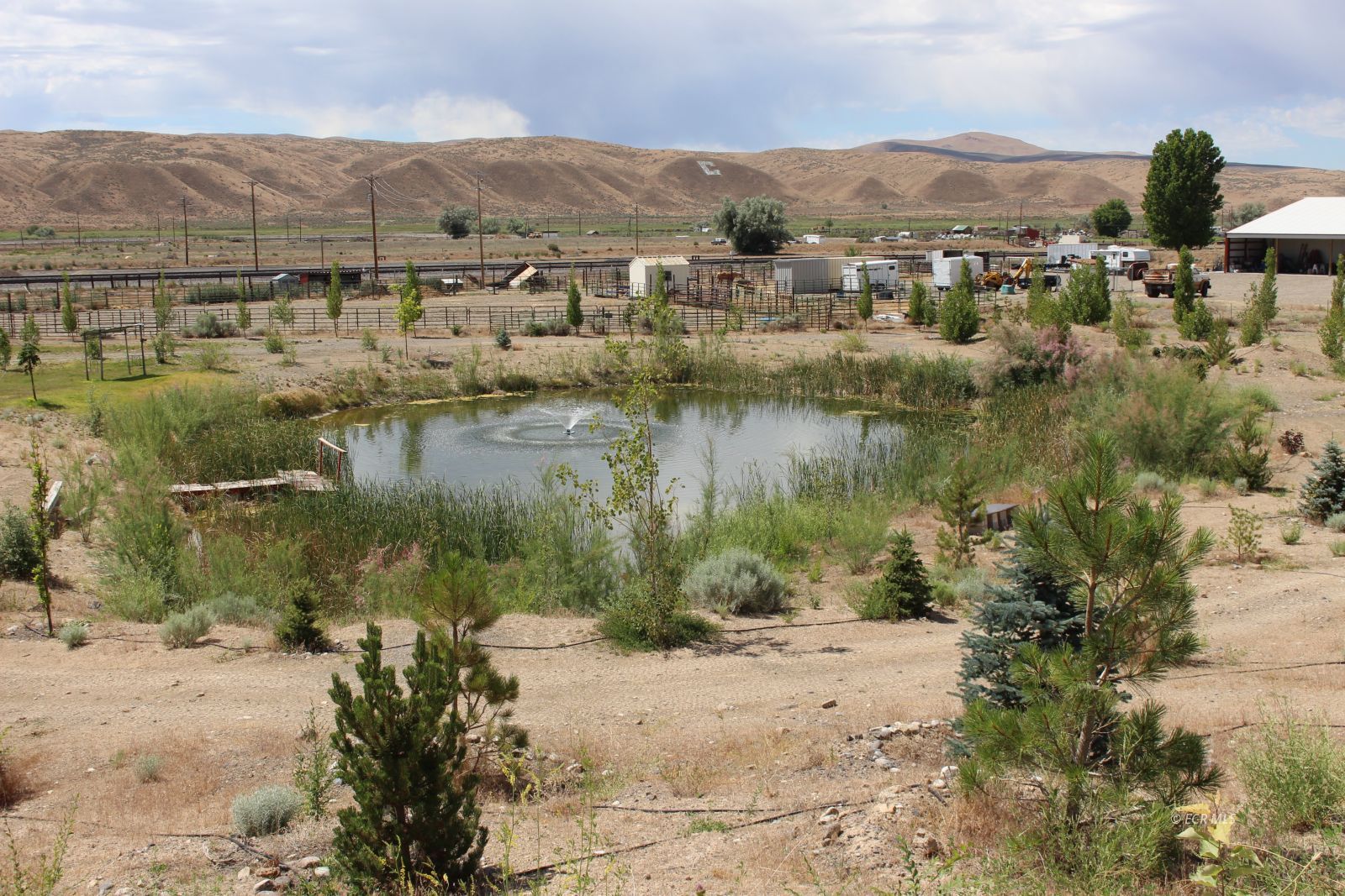 Rural landscape with a pond and fountain, fenced pens, and dry hills.