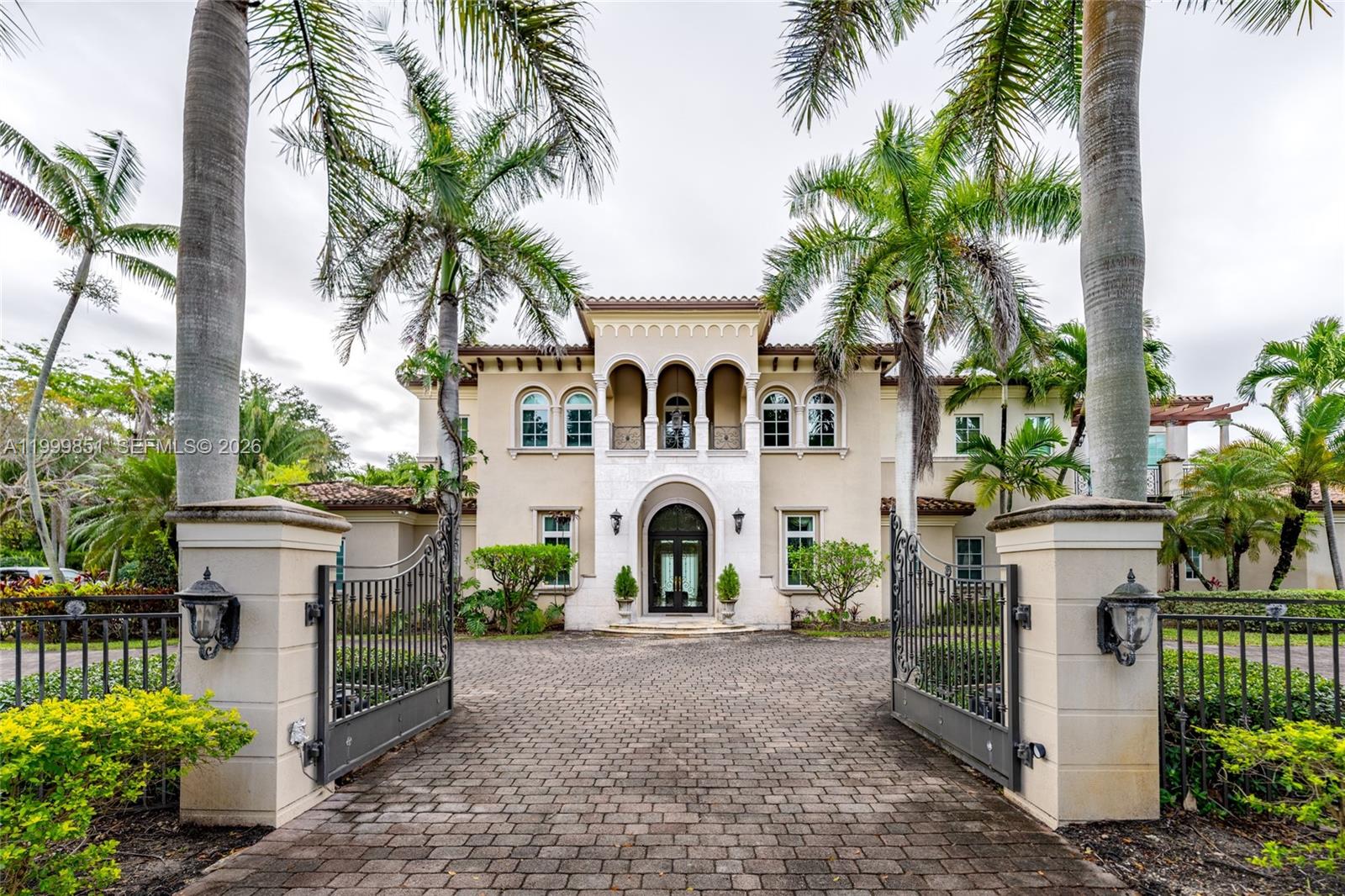 A statement of timeless elegance and architectural grandeur, this Mediterranean-inspired estate welcomes you through a gated entrance framed by majestic royal palms and a sweeping circular driveway. The striking façade showcases symmetrical design, arched windows, and a second-story balcony that sets an impressive tone of refinement.Step inside to a breathtaking double-height foyer featuring a dramatic curved staircase with wrought-iron detailing, gleaming marble floors, and a grand arched entry door that fills the space with natural light. Every element reflects meticulous craftsmanship and a sense of arrival.The main living areas are designed for both comfort and sophisticated entertaining, highlighted by soaring ceilings, expansive open spaces, and elegant archways that seamlessly connect each room. The formal living room exudes warmth and luxury, complemented by a second-floor balcony overlook that enhances the home’s open, airy ambiance.Architectural details such as custom iron railings, columns, and curated finishes elevate the interiors, while large windows and glass doors invite in natural light and offer tranquil views of the lush surroundings.This residence embodies the essence of South Florida luxury living—grand yet inviting, refined yet comfortable—offering a perfect setting for upscale entertaining and everyday elegance.