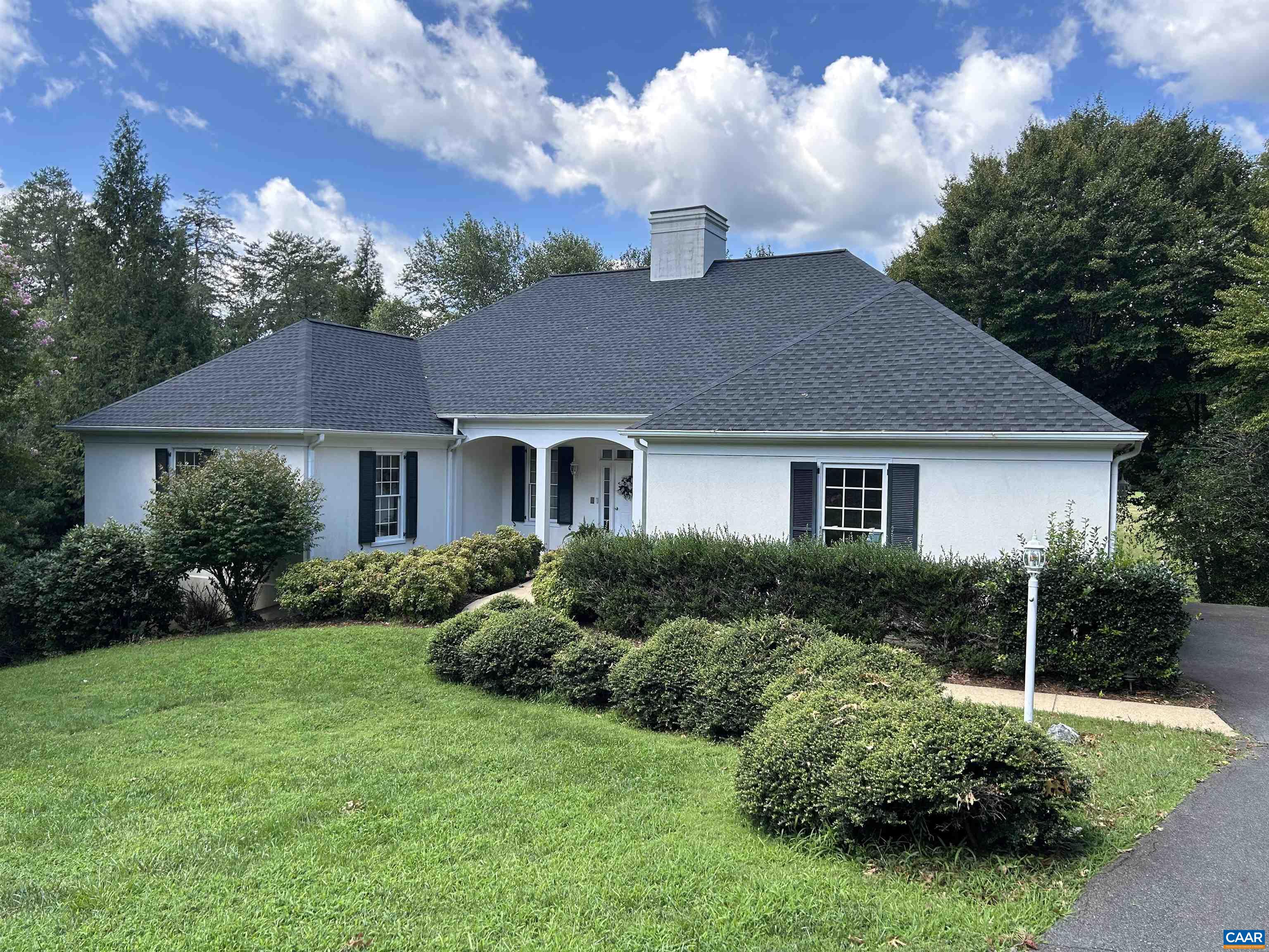 White single-story home with a broad gray roof, covered entry, and manicured shrubs along the lawn.