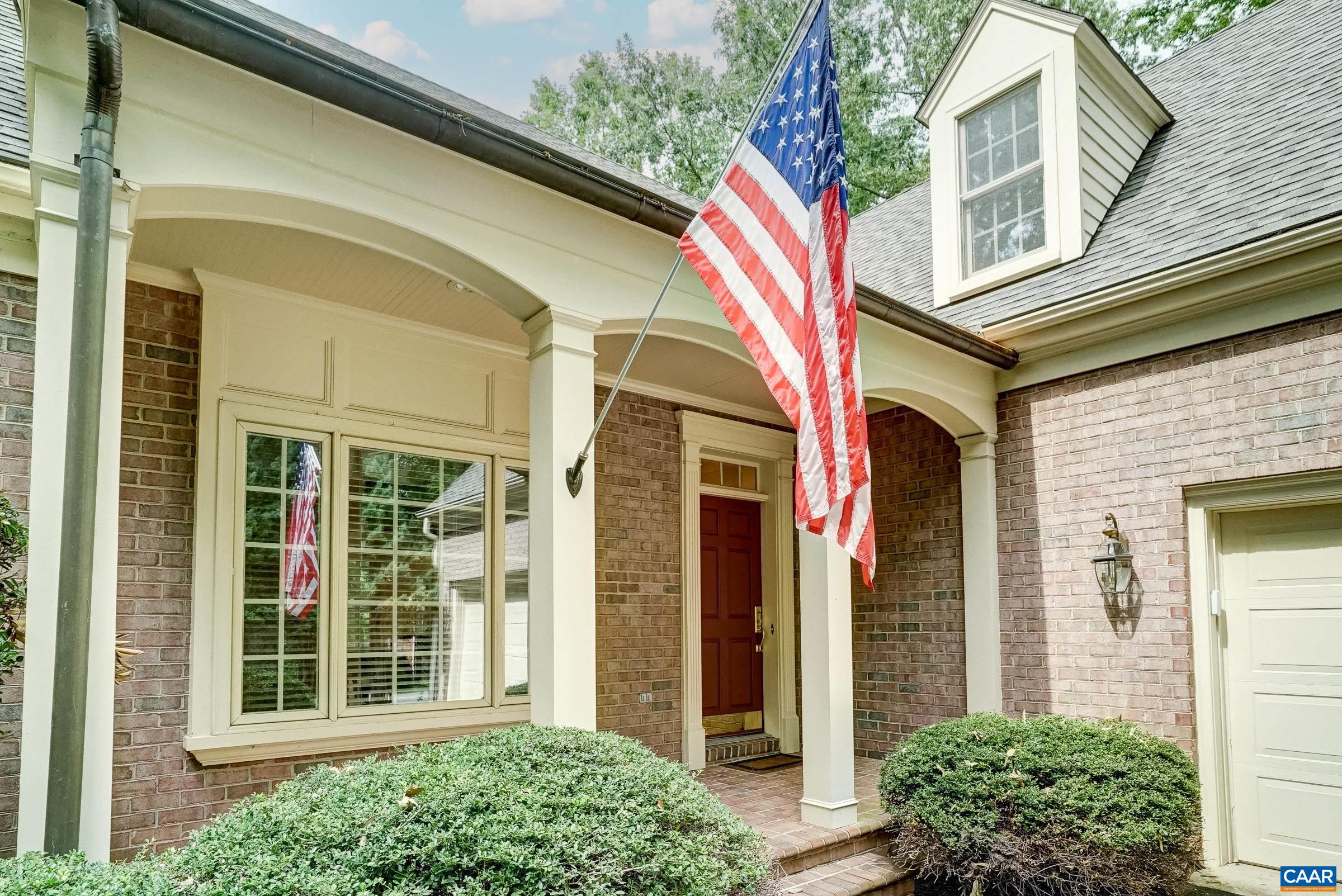 Brick home entrance with a covered porch, red door, American flag, and trimmed shrubs.