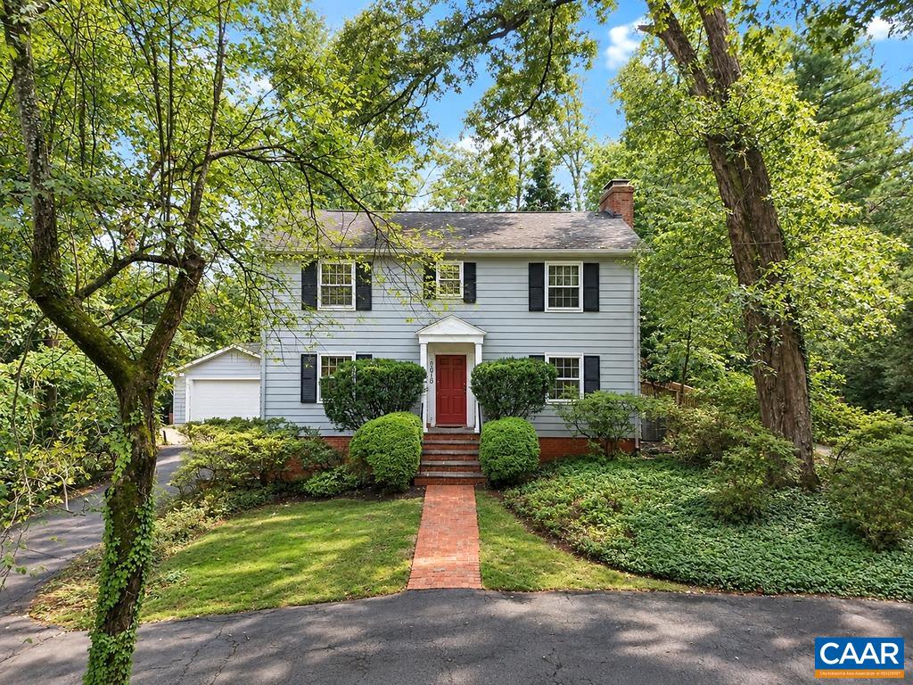 Two-story blue house with black shutters, a red door, and mature trees framing the front yard.