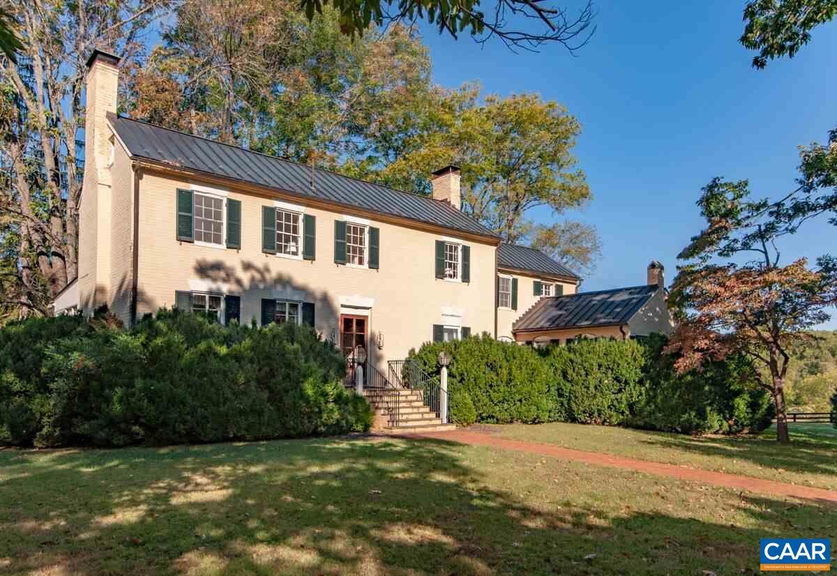 Cream-brick house with dark metal roofs and green shutters behind hedges and a lawn.
